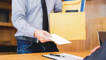 An employee, wearing a shirt and tie, extends a letter of resignation to their boss while holding a box of documents.