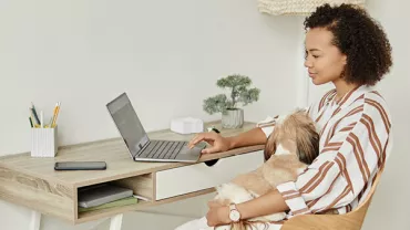 Persoon zit aan een bureau en gebruikt een laptop met een hond op schoot.